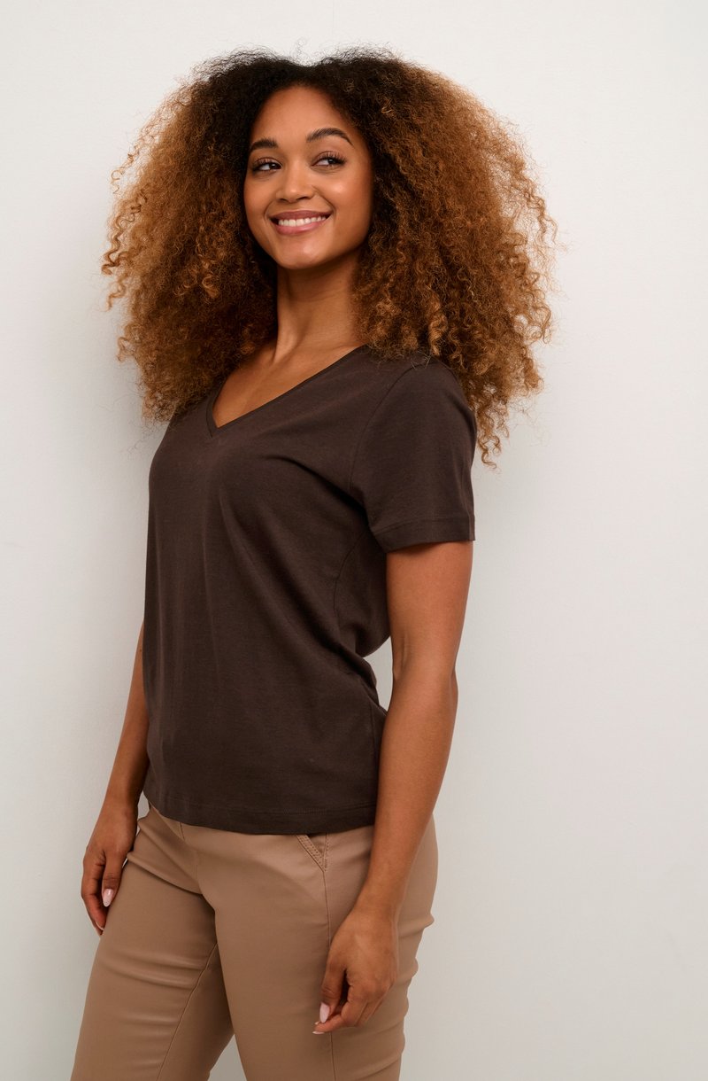 Camiseta de algodón marrón con escote en V y mangas cortas, combinada con pantalones beige claros. El modelo está de lado, mostrando un ajuste relajado.