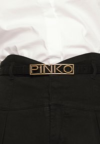 Svarta byxor har en guld "PINKO" bältesspänne, med en slät yta och skräddarsydd passform. Bältet är fäst i midjan.