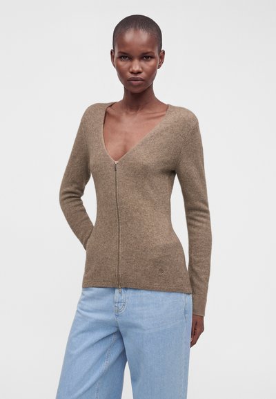 Femme portant un pull marron ajusté zippé avec un col en V profond et un jean bleu clair, debout avec une main derrière le dos.