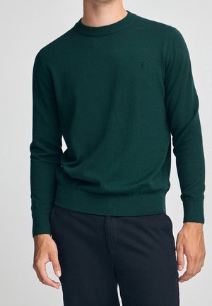 Mężczyzna w ciemnozielonym swetrze z okrągłym dekoltem z małym haftowanym logo na piersi i czarnych spodniach, stojący na jasnym tle.