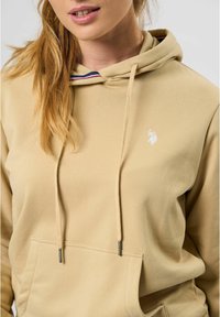 Beige hettegenser med snorhette, frontlomme og hvit logo på brystet. Har stripete detaljer inni hetten. Myk tekstur.