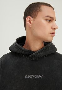 Mladić s kratkom kosom koji nosi crnu majicu s kapuljačom na kojoj piše "URTRN", gleda udesno na jednostavnoj svijetloj pozadini.