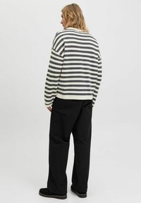 Striped, loose-fitting sweater in gray and white with dropped shoulders, paired with wide black trousers and black slip-on shoes.