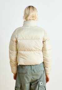 Veste courte matelassée en beige clair, avec un col montant, un design matelassé et des poignets élastiques. Portée avec un pantalon cargo vert.
