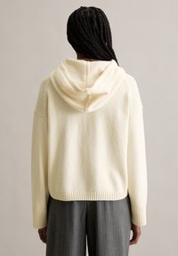 Persona con i capelli intrecciati che indossa una felpa con cappuccio beige lavorata a maglia e pantaloni grigi a righe sottili, vista da dietro su uno sfondo semplice.