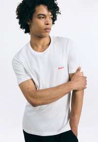 Tânăr cu păr creț, îmbrăcat cu un tricou alb cu logo-ul roșu „HUGO”, ținându-se de brațul superior stâng, privind spre dreapta, pe un fundal simplu.