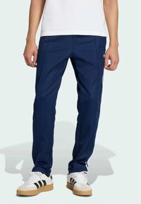 Ναυτικά αθλητικά παντελόνια από ελαφρύ ύφασμα με πλευρικές ρίγες και στενές γραμμές, συνδυασμένα με λευκά αθλητικά παπούτσια με μαύρες ρίγες.