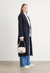 Abrigo largo en azul marino con solapas anchas, puños forrados y un bolso crema. La modelo lleva un suéter crema y jeans en azul claro con zapatillas blancas.