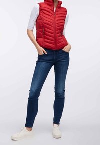 Rood donzen vest met ritssluiting aan de voorkant, horizontaal doorgestikt patroon en twee zijzakken. Gecombineerd met blauwe strakke jeans en witte sneakers.