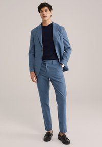Hele sinine kohandatud ülikond, millel on tekstuuriga muster, sobitatud tume navy crew neck särke ja mustade mokassiinidega. Puhas, modernne disain.