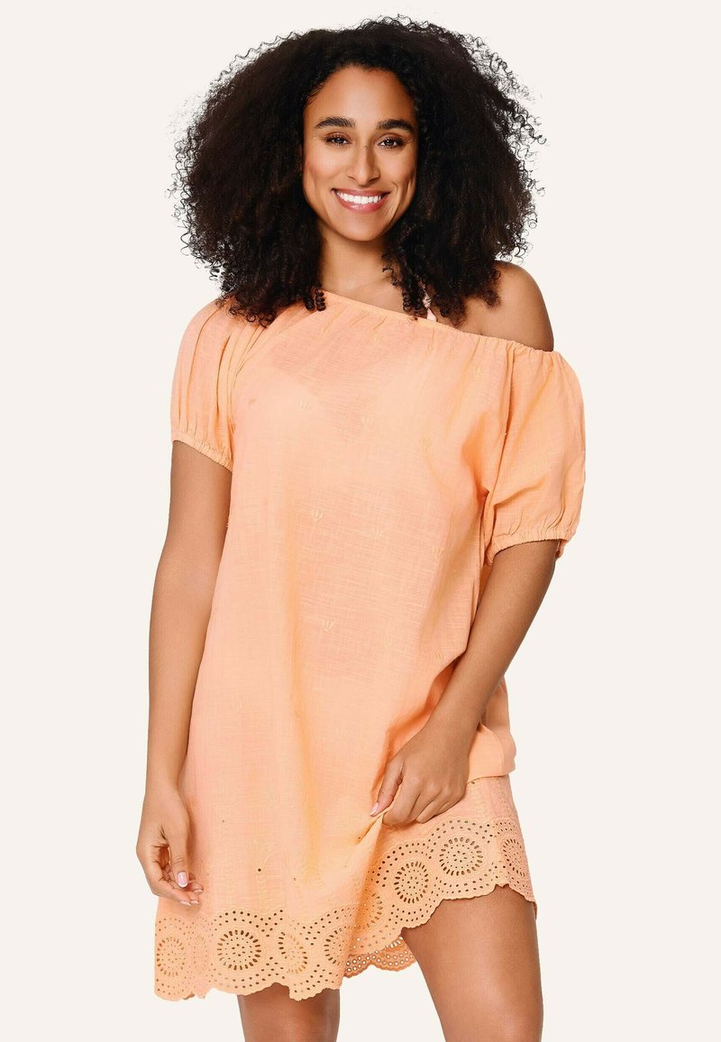Femme souriante aux cheveux bouclés portant une robe pêche à épaules dénudées avec un ourlet en dentelle ajourée, debout devant un fond clair uni.