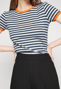 Kobieta w granatowo-białej koszulce w paski z pomarańczowym wykończeniem i logo, w połączeniu z czarnymi spodniami z wysokim stanem, ręce na biodrach.