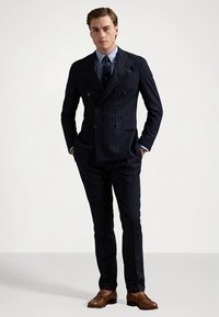 Marineblauw pak met krijtstreep, bestaande uit een dubbelzijdig jasje en slim-fit broek. Gecombineerd met een lichtblauw overhemd en bruine leren schoenen.