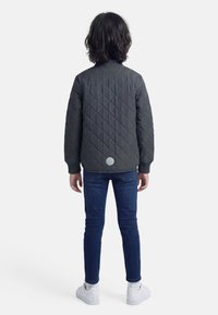 Jauns cilvēks ar plecu garuma tumšiem matiem, valkājot tumšu polsterētu jaku, zilas džinsas, baltas zeķes un baltas sporta kurpes, stāv ar muguru pret skatītāju.