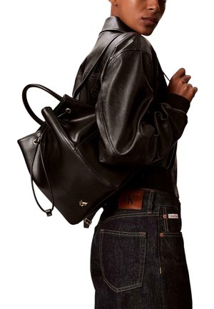 Sac à dos noir en cuir avec cordon de serrage et ferrures contrastantes, porté à l'épaule. Le modèle porte une veste bomber noire et un jean en denim.