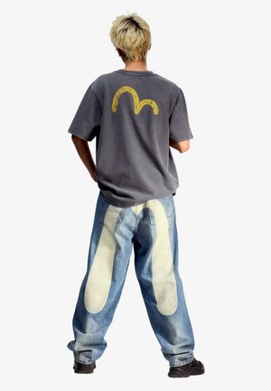 Persona con capelli biondi corti che indossa una maglietta grigio scuro con un disegno giallo e jeans blu larghi con grandi impronte bianche sul retro.