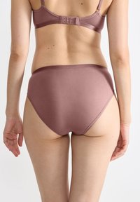 Vrouw in een mauve bh en bijpassende high-waist onderbroek gezien vanuit de achterkant tegen een plain witte achtergrond.