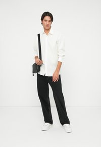 Cămașă albă cu mâneci lungi și nasturi, blugi negri evazați, geantă crossbody neagră și adidași albi, stând pe un fundal simplu.