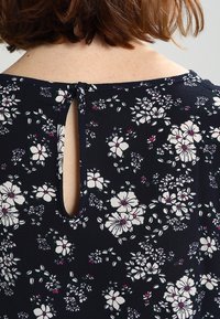 Blusa floreale blu navy con scollatura rotonda e apertura a chiave sul retro, decorata con fiori bianchi e rosa su un tessuto testurizzato.