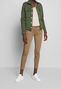 Kvinna iklädd en grön knapplös jacka, beige V-ringad topp, tan slim-fit byxor och vita sneakers som står mot en enkel grå bakgrund.