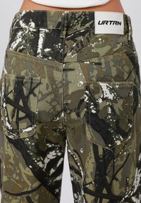 Pantaloni con motivo camouflage in diverse tonalità di verde e nero. Presentano due tasche posteriori e un'etichetta bianca con il marchio "URTRN" in vita.