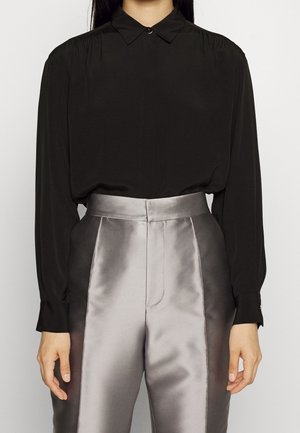 Personne portant un chemisier noir à manches longues boutonné, rentré dans un pantalon argenté-gris brillant taille haute avec poches latérales.