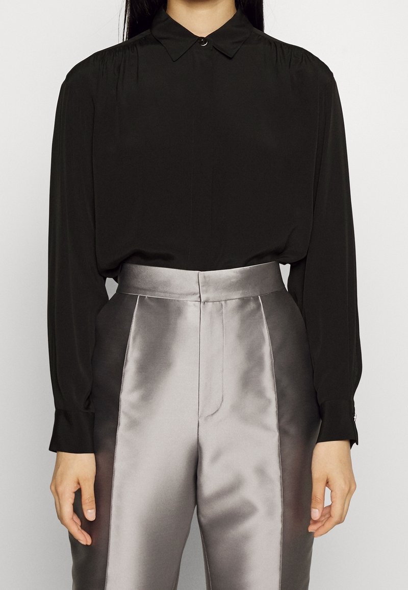 Personne portant un chemisier noir à manches longues boutonné, rentré dans un pantalon argenté-gris brillant taille haute avec poches latérales.