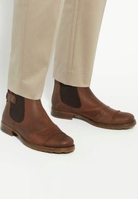 Brown leather ankle boots with elastic side panels, rounded toe, cap toe design, and a low stacked heel. Tan trousers visible above the boots.