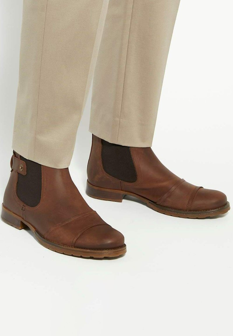Brown leather ankle boots with elastic side panels, rounded toe, cap toe design, and a low stacked heel. Tan trousers visible above the boots.
