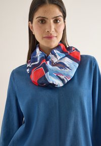 Femme aux longs cheveux bruns portant un haut bleu et une écharpe multicolore avec des motifs abstraits rouges, bleus et blancs autour du cou.