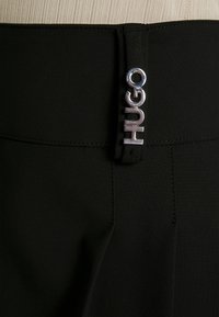 Pantaloni neri con tessuto testurizzato, una vita ampia e un dettaglio in argento sul davanti. Design sartoriale con cuciture a vista.