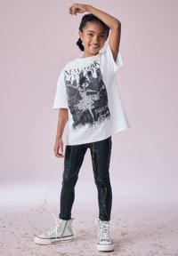 Maglietta bianca con grafica di balletto nera, abbinata a leggings neri lucidi e sneaker alte argento. Lo sfondo presenta un rosa tenue.
