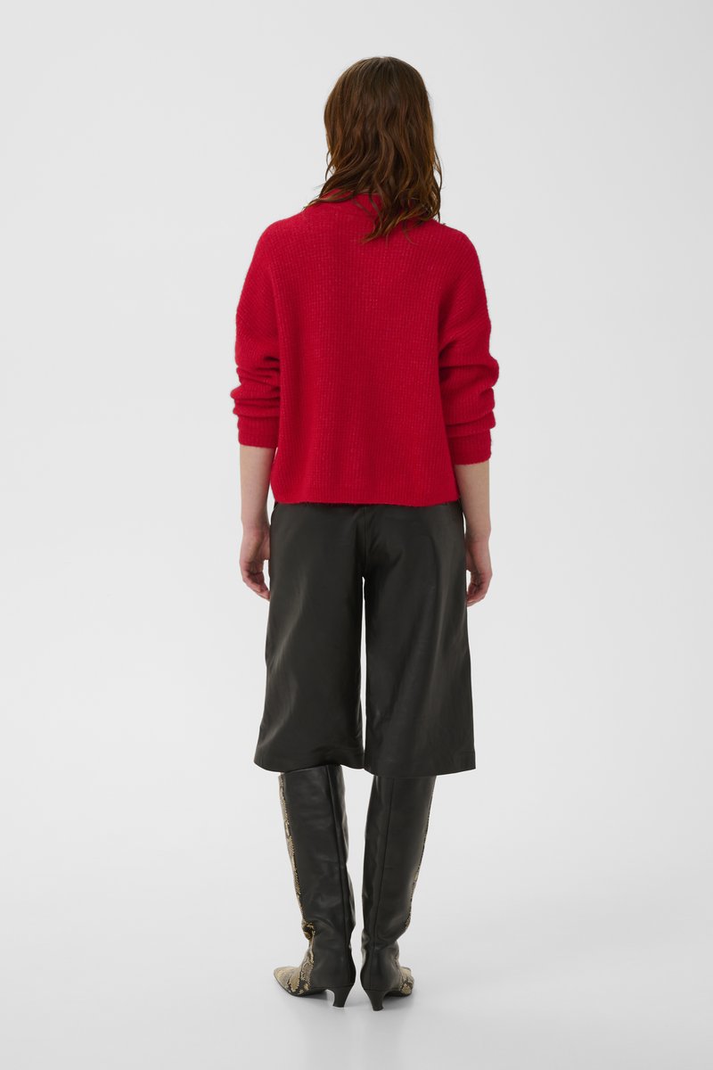 Femme aux cheveux bruns ondulés portant un pull rouge en maille, un pantalon en cuir noir à jambes larges et des bottes noires montantes avec un motif peau de serpent.