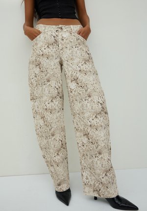 Kobieta nosząca beżowe spodnie z wysokim stanem i szerokimi nogawkami w cętkowany wzór, czarne szpilki z noskiem w szpic oraz krótki czarny top zapinany na guziki.