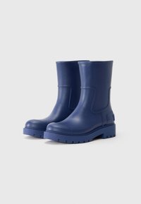 Bottes de cheville en caoutchouc bleu avec une texture lisse, bout rond et semelle épaisse, dotées de panneaux latéraux pour un enfilage facile et de détails de couture minimaux.