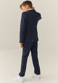 Navyblauw geruit pak met een getailleerde pasvorm, voorzien van een enkelgeknoopte blazer en bijpassende broek. Witte sneakers maken de look compleet.