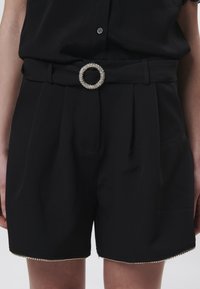 Pantalones cortos negros de cintura alta con un diseño plisado, que cuentan con un cinturón decorativo con una hebilla de rhinestone circular y un dobladillo adornado.