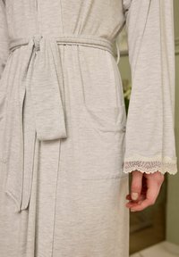 Robe grigio chiaro con cintura, dotata di maniche lunghe e rifiniture in pizzo sui polsini. Include una tasca frontale funzionale. Tessuto morbido.