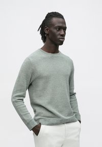 Világoszöld kötött pulóver texturált mintával, kerek nyakkivágással és bordázott mandzsettákkal, világos színű nadrág fölött viselve.