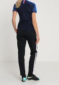 Persona de pie, vistiendo una camisa azul marino con una franja roja, blanca y azul, pantalones de chándal negros con una franja blanca y zapatillas negras con acentos en verde neón.