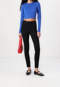 Blå kortärmad topp med långa ärmar, svarta högmidjade skinny jeans, röd handväska och svarta platta skor med ögla-accenter.