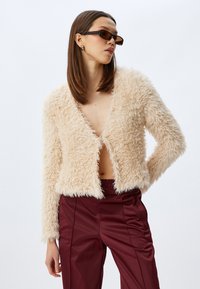 Kürzerer Kunstfelljacke in hellbeige, mit tiefem V-Ausschnitt, langen Ärmeln und einer weichen, strukturierten Oberfläche. Kombiniert mit burgunderfarbener Hose.