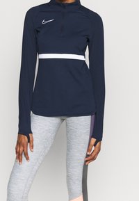 Maglietta atletica a maniche lunghe color navy con apertura a mezzo zip, striscia orizzontale bianca e logo Nike; abbinata a leggings grigio melange. Tessuto morbido e aderente.