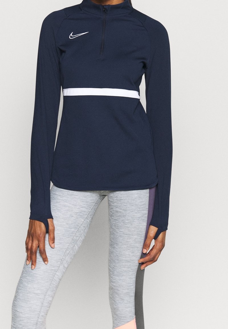 Maglietta atletica a maniche lunghe color navy con apertura a mezzo zip, striscia orizzontale bianca e logo Nike; abbinata a leggings grigio melange. Tessuto morbido e aderente.