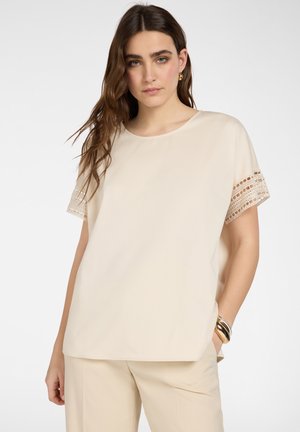 Giovane donna che indossa una camicetta crema a maniche corte con dettagli in pizzo sulle maniche e pantaloni abbinati, accessoriata con braccialetti e orecchini dorati.