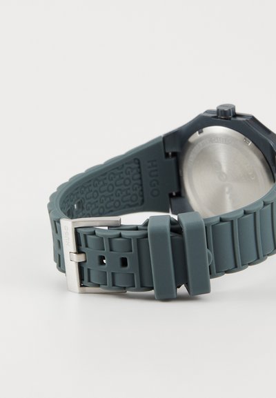 Montre Hugo avec un bracelet en silicone gris foncé arborant un motif en grille et une boucle en métal argenté, mettant en avant un design minimaliste et une texture lisse.
