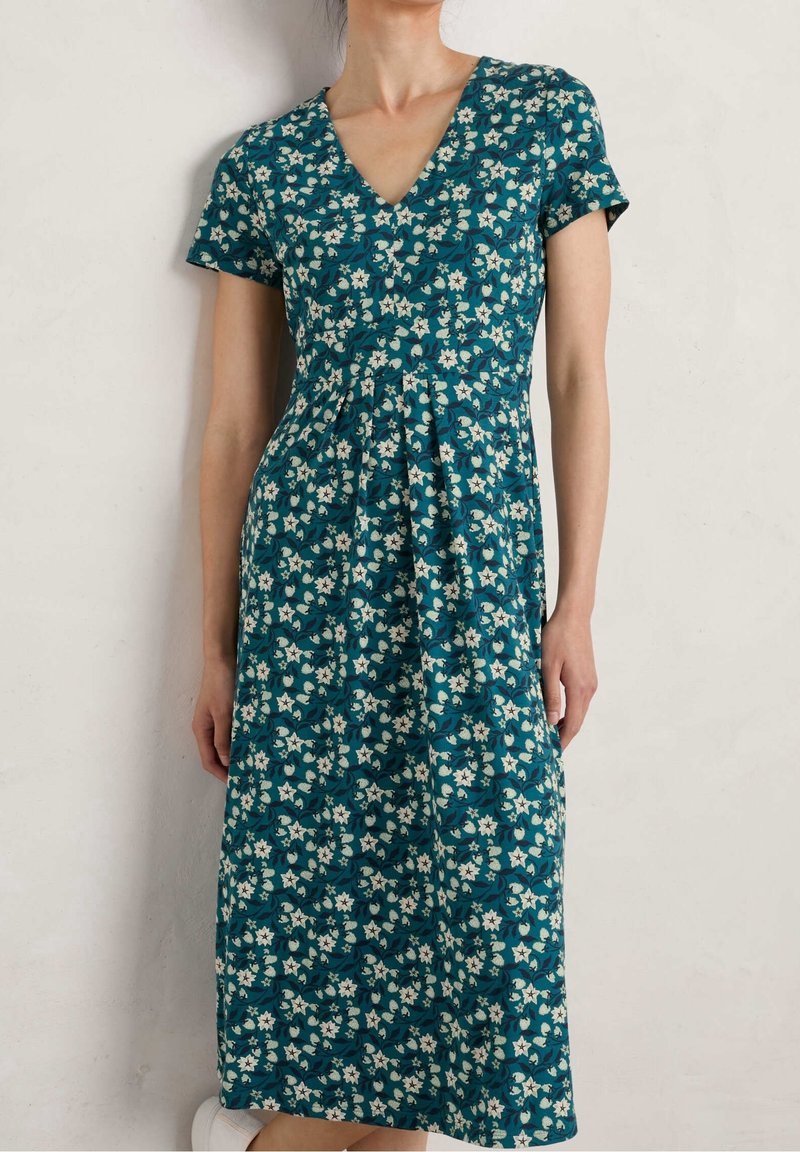 Femme portant une robe midi bleu sarcelle à manches courtes avec un motif floral blanc, debout contre un mur clair uni.
