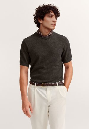 Man with curly hair wearing dark textured knit short-sleeve shirt, striped neck scarf, white pants, and braided brown belt, looking right.