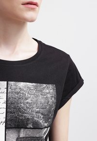 Camiseta de algodón negra con mangas cortas enrolladas, que presenta un diseño gráfico de texturas en blanco y negro y elementos escritos a mano en el frontal.