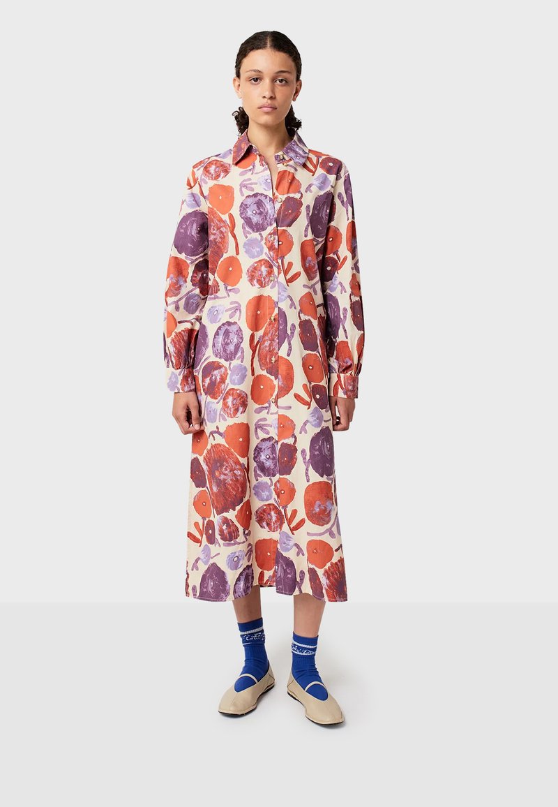 Kobieta stoi w długiej, zapinanej sukience w czerwone i purpurowe kwiatowe wzory, w niebieskich skarpetach w wzory oraz beżowych płaskich butach na białym tle.